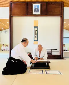Two men in martial arts uniforms sign certificates in a dojo with seated practitioners.