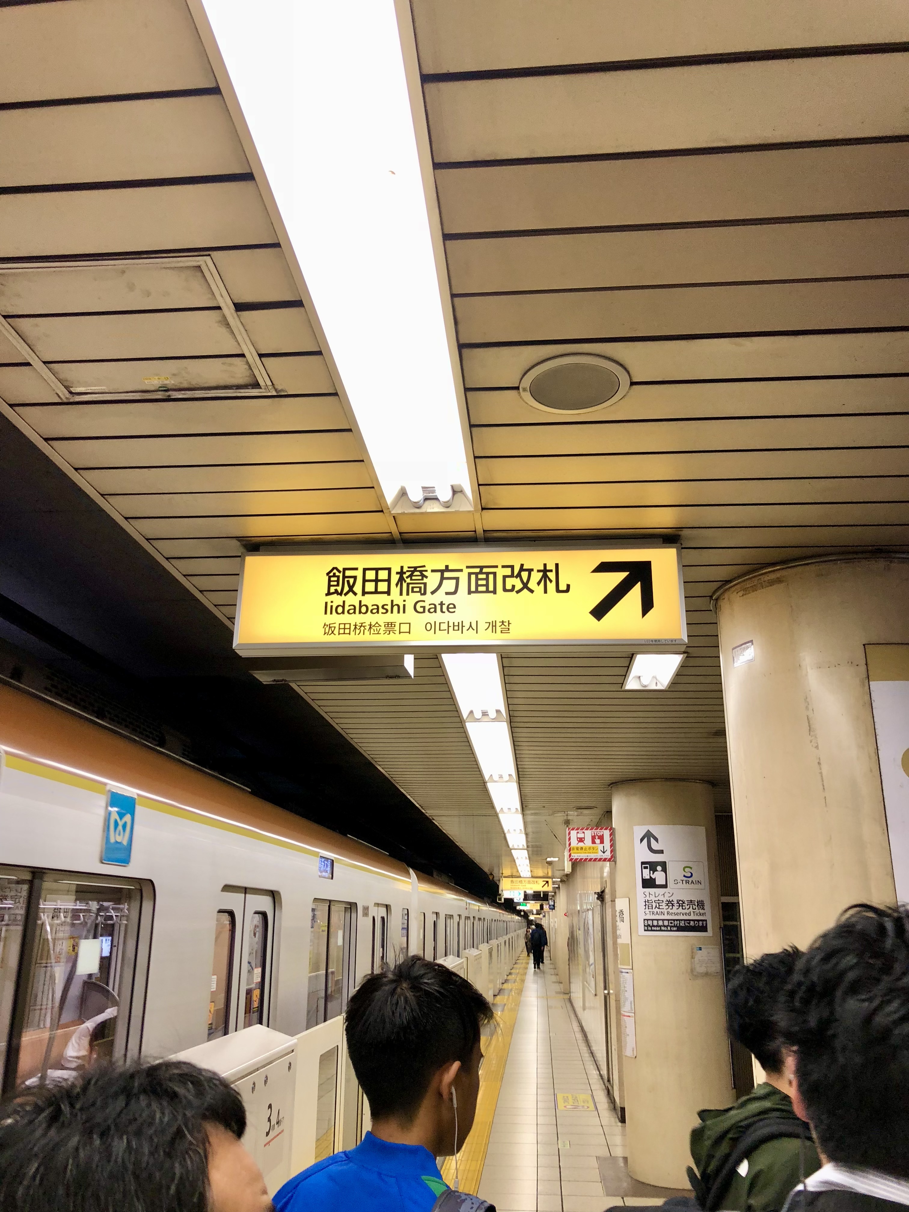 Subway platform with train on left, passengers waiting, and overhead Iidabashi Gate sign in four languages.