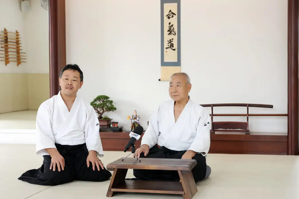 Two men in martial arts uniforms seated on the floor with a microphone and dojo decor behind.