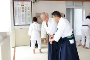 Two men practice a wrist technique in a bright dojo with other martial artists training nearby.
