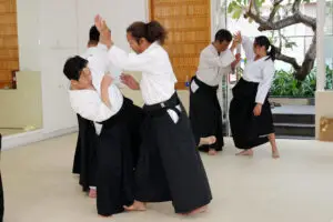 People practicing paired martial arts techniques wearing white tops and black hakama in a dojo.