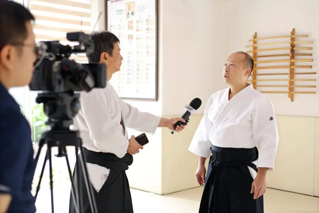 Martial arts instructor in white gi and black belt interviewed in dojo with wooden weapons on wall.