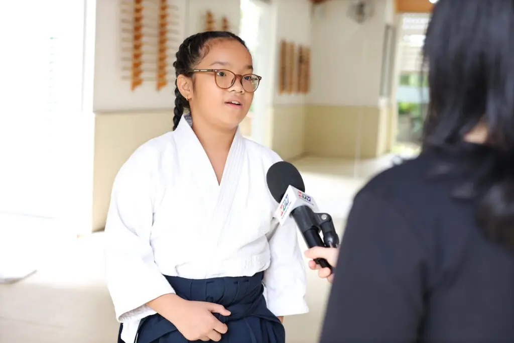 Young person in white martial arts uniform and glasses being interviewed indoors.