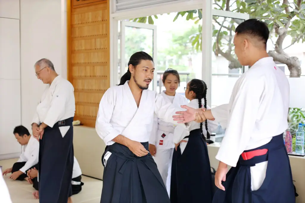 People wearing martial arts uniforms practicing techniques and preparing indoors near open windows.