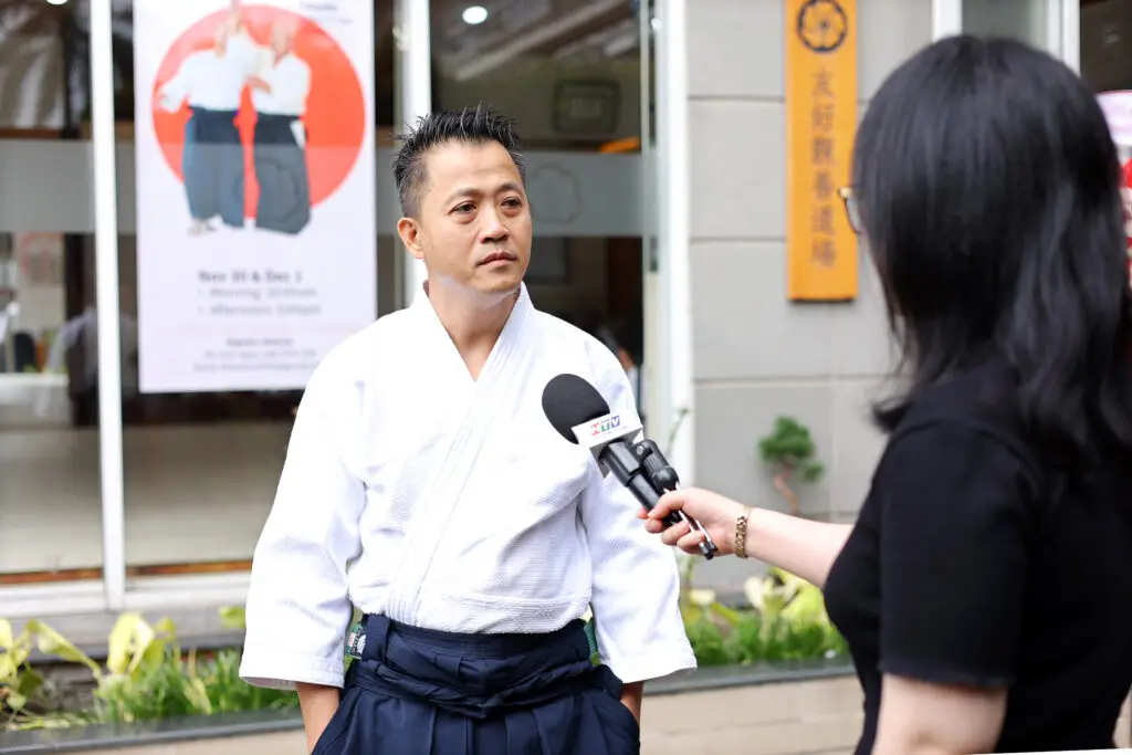 Man in martial arts uniform being interviewed with microphone outside a building.