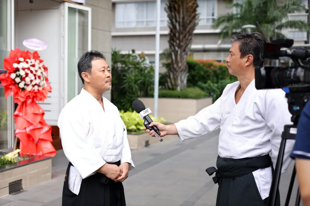 Two men in martial arts uniforms conduct an outdoor interview with a camera nearby.