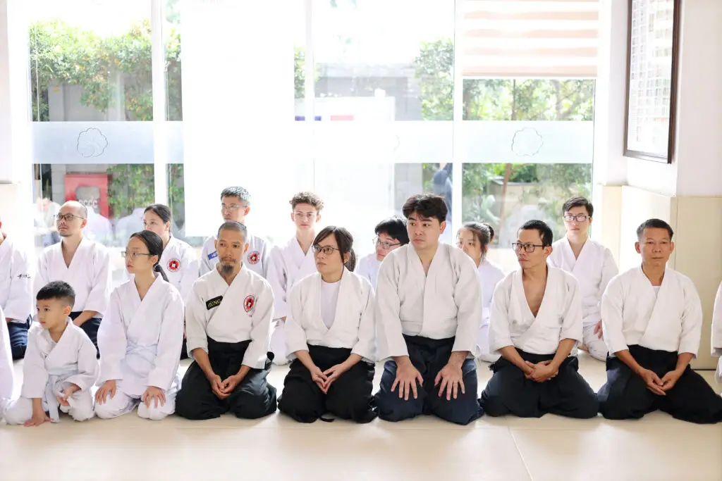 Group of people in martial arts uniforms seated kneeling indoors near large windows with greenery outside.