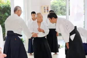 Martial arts instructor demonstrates technique to students dressed in traditional attire inside bright dojo.