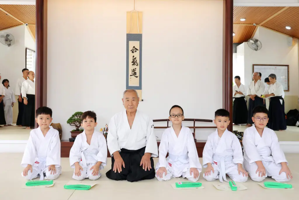 Martial arts instructor and five children kneeling with certificates and green belts inside dojo.