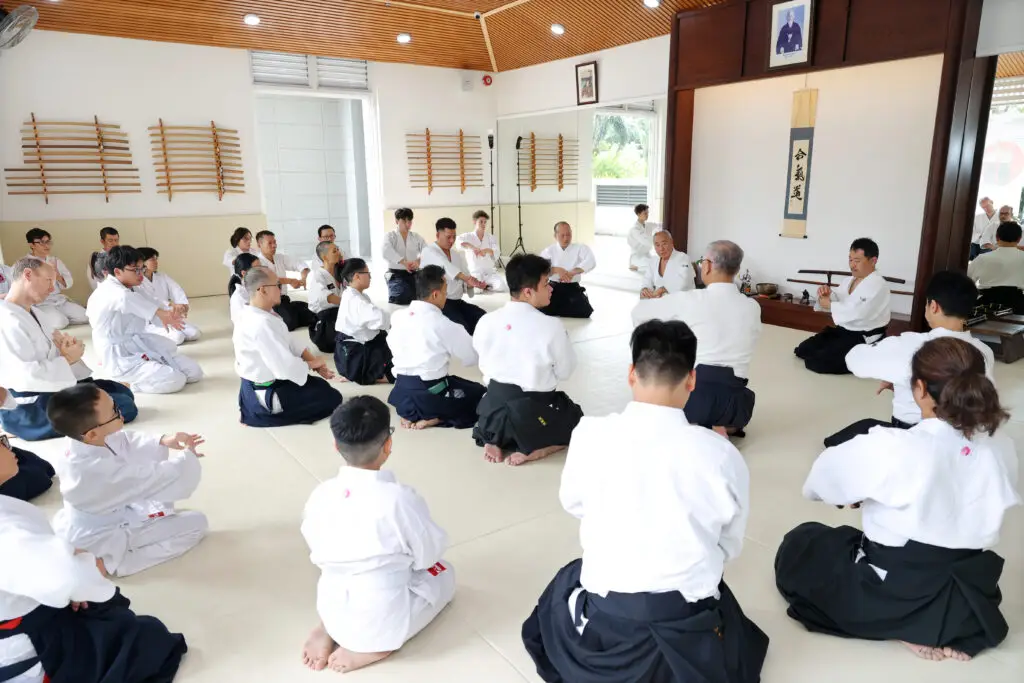 Martial arts class with students seated on mats facing an instructor in a dojo interior.