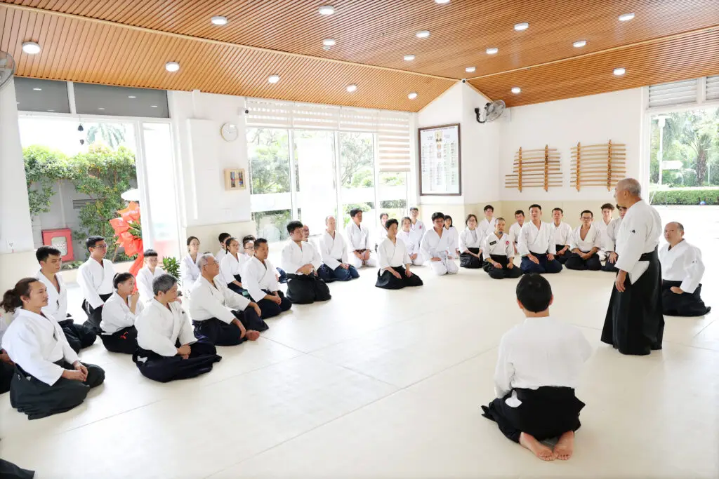 Martial arts class with students seated on mat listening to standing instructor in bright dojo.