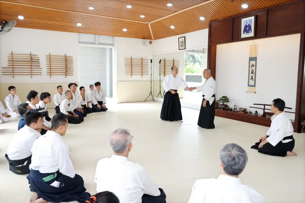 Martial arts class with practitioners in white uniforms watching two instructors demonstrating a technique in dojo.