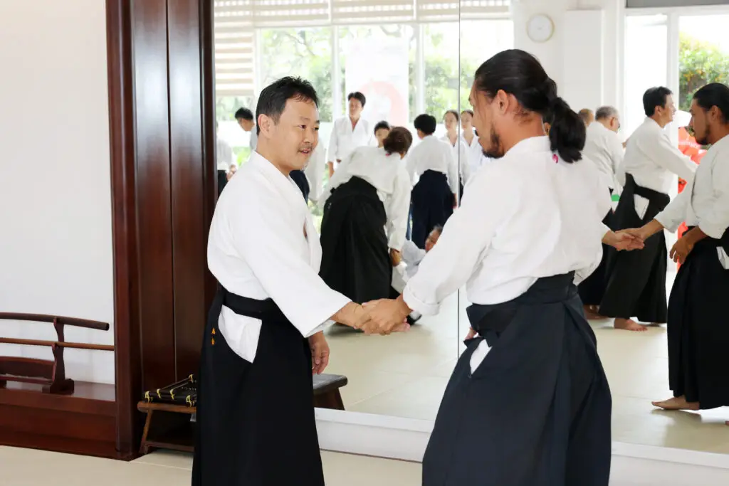 Two men in martial arts uniforms shaking hands in a dojo with others practicing in the background.