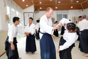 People practicing Aikido techniques in a brightly lit dojo with wooden ceiling and windows.