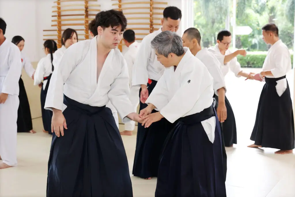 People practicing paired hand techniques in white jackets and black hakama pants in a dojo.