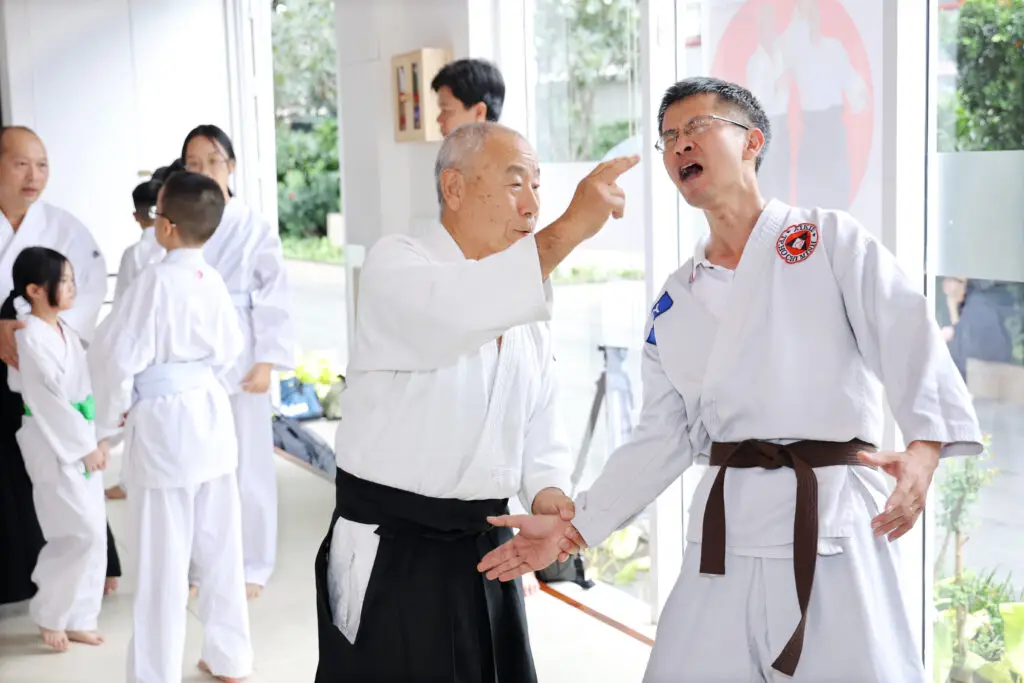Martial arts instructor demonstrates a technique on a student in a dojo with others present.