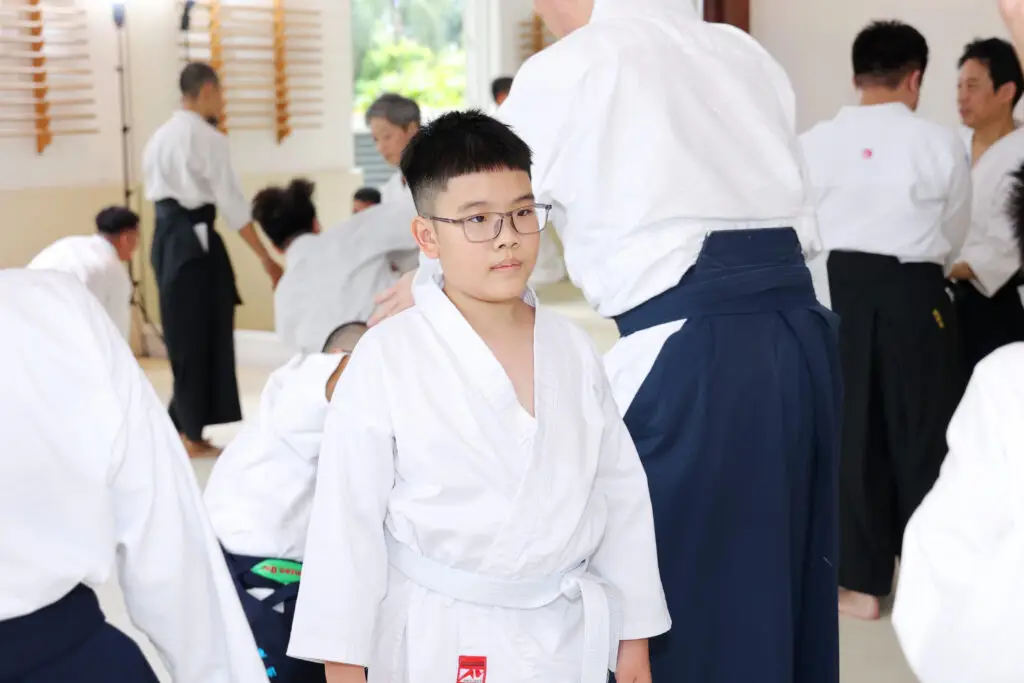 Boy with glasses in white martial arts uniform with white belt inside a dojo class.