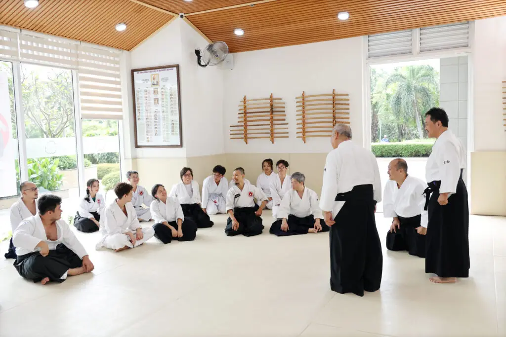 Martial arts class with participants in white uniforms seated and instructors standing in dojo.