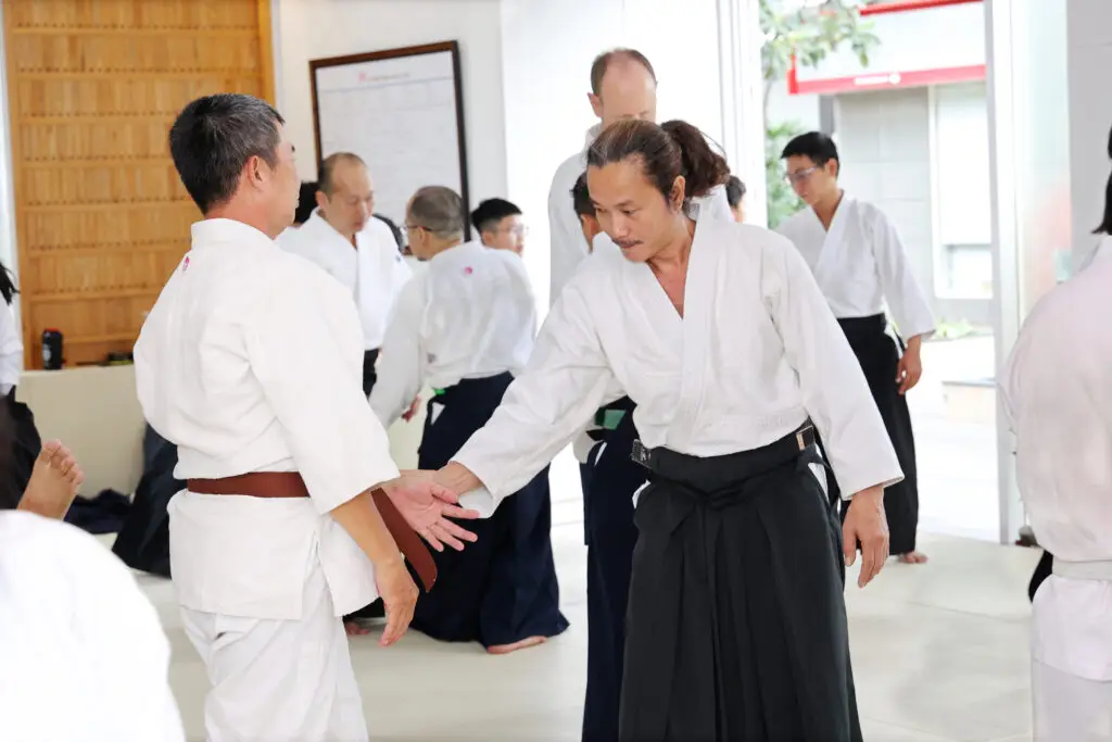 Martial arts practitioners in white uniforms train hand grip techniques in a bright indoor dojo.