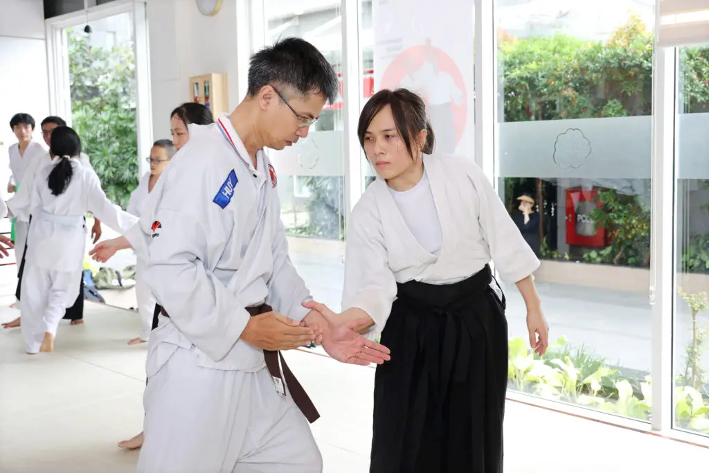 Two individuals in martial arts uniforms practicing a technique indoors near large windows.