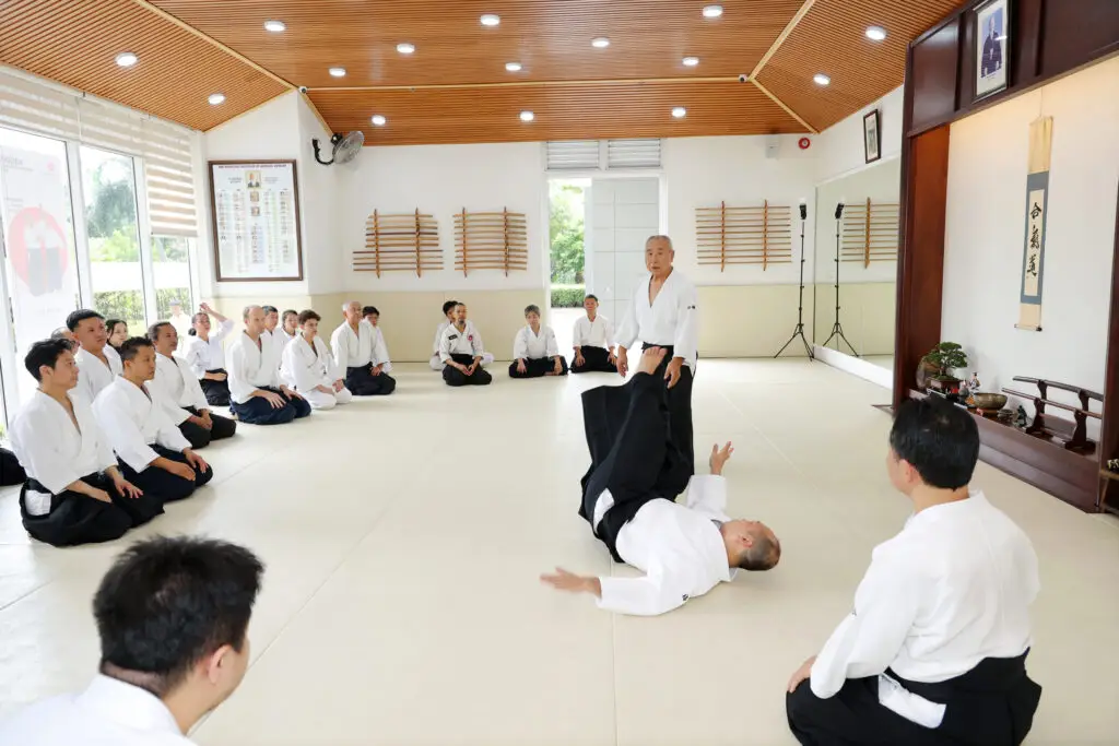 Martial arts class in dojo with students seated watching technique demonstration on mats.