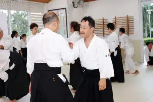 People practicing paired martial arts techniques in a bright dojo with martial arts equipment.