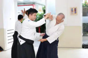 Two men in martial arts uniforms practicing Aikido techniques indoors with others training in background