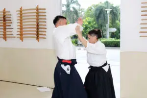 Two martial artists in traditional uniforms practice techniques inside a dojo near an open doorway.