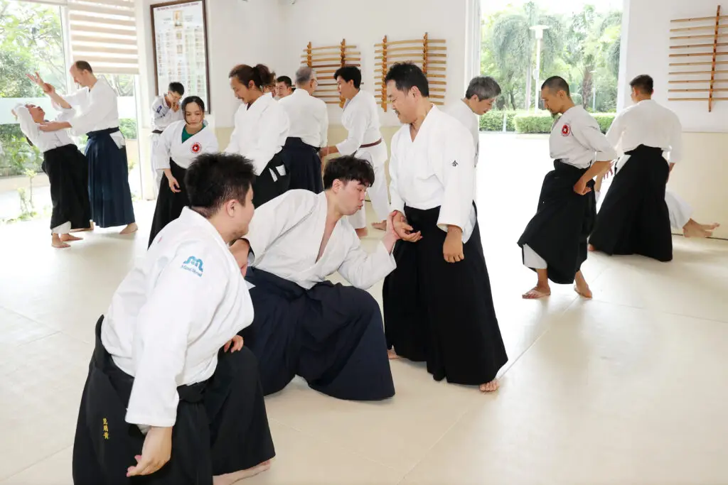 Group practicing Aikido techniques in a bright dojo with wooden training weapons on walls.