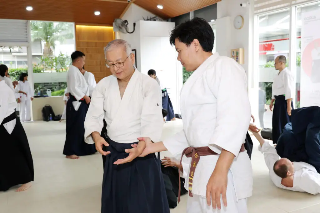 Two men demonstrating a wrist technique in martial arts class with other students practicing.