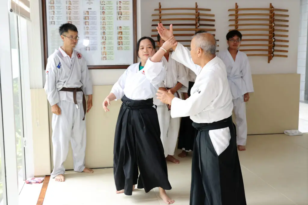 Martial arts instructor demonstrates technique on female student with others observing indoors.