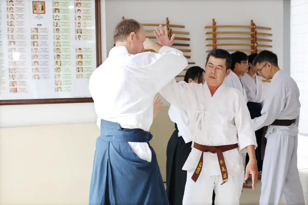 Martial artists practicing Aikido techniques in a dojo with wooden weapons and a history chart.