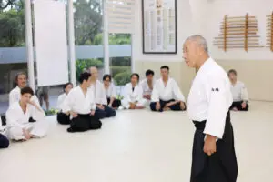 Martial arts instructor speaking to seated students in a dojo with weapons on the wall.