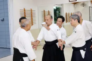Six martial artists in traditional uniforms practice a joint lock technique in a dojo.