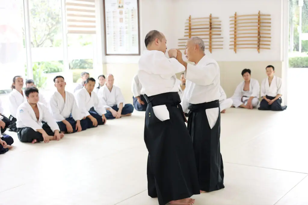 Martial arts instructors demonstrate technique to seated students in a bright dojo with weapon racks.