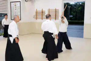 Four people practicing martial arts with wooden weapons in a dojo with swords on wall.