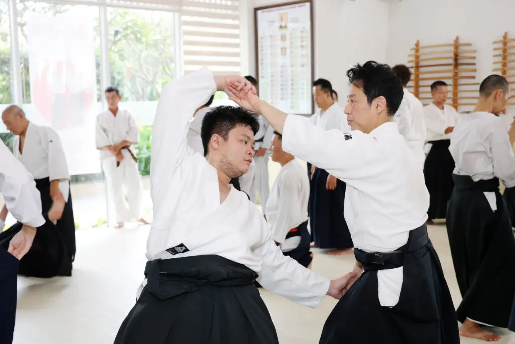 Two martial artists practicing a technique indoors wearing white uniforms and black pants.