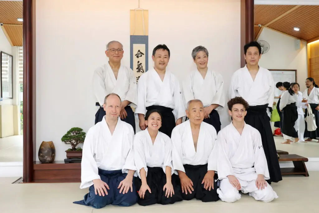 Eight martial artists in white uniforms pose inside a dojo with bonsai and calligraphy.