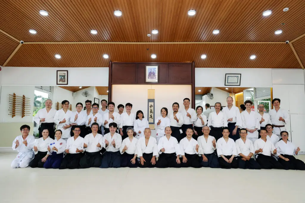 Martial arts group in white uniforms posing in dojo with wooden ceiling and calligraphy display.