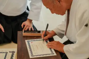 Older man in martial arts uniform signing a certificate on a wooden table with another person nearby.