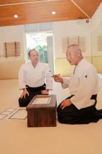 Two martial artists in a dojo kneeling with certificates and a wooden table between them.