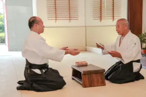 Two men in martial arts attire kneeling on a dojo mat exchanging a certificate over a wooden table.