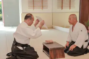Two men in martial arts uniforms seated on the floor inside a dojo with a small wooden table.