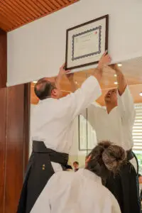 Two martial artists hang a framed certificate on a dojo wall with mirrors.